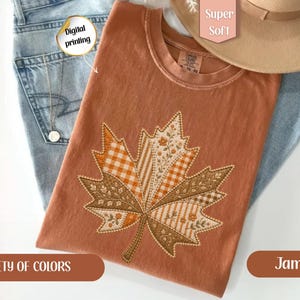 May include: A rust-colored t-shirt featuring a large embroidered maple leaf design composed of various patterned fabric sections, including orange gingham, floral prints, and striped textures, layered over light blue denim jeans and a tan hat.