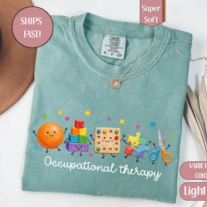 May include: A light green t-shirt featuring a graphic of cartoon occupational therapy tools including an orange ball, colorful stacking blocks, a pegboard, a puzzle piece, and scissors, with the text Occupational therapy printed below.