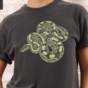 Peut inclure: T-shirt gris foncé avec une illustration de serpent crème et vert. La marque Comfort Colors et la couleur « Pepper » sont affichées en haut à gauche. Les manches sont retroussées.