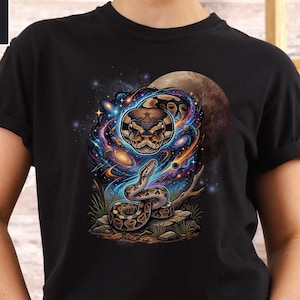 May include: Black t-shirt with a vibrant graphic of a snake set against a cosmic backdrop. The design includes a detailed snake, galaxies, stars, and a moon. The shirt is made of a soft material, perfect for casual wear.