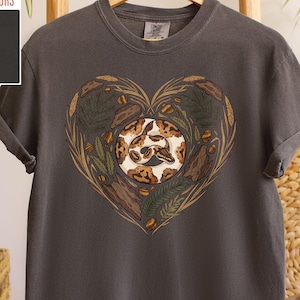 Op de afbeelding: Pepperkleurig T-shirt met een hartvormig ontwerp met een slangenillustratie. Het hart is gevuld met bladeren, hout en eikels. Het shirt is gemaakt van een zacht, comfortabel materiaal.