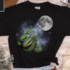 May include: Black t-shirt featuring a graphic of three green snakes with a full moon and a starry background. The shirt is made of a soft material and is a Comfort Colors brand.