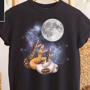 Op de afbeelding: Zwart T-shirt met een afbeelding van drie slangen met open monden, onder een volle maan en een sterrenstelsel op de achtergrond. De slangen zijn bruin, geel en wit. Het shirt hangt aan een houten hanger.