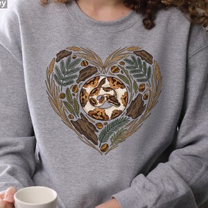 Puede incluir: Sudadera Sport Grey con un diseño en forma de corazón. El diseño presenta una serpiente en el centro, rodeada de hojas, corteza y nueces. La serpiente tiene marcas marrones y beige. La sudadera es de color gris sólido.