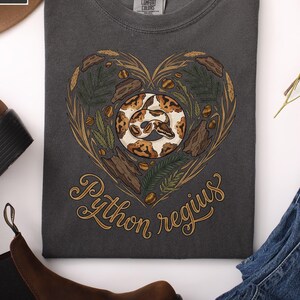 Op de afbeelding: Donkergrijze t-shirt met een hartvormig ontwerp met een slang, bladeren en de woorden "Python regius" in gouden letters. De t-shirt is opgevouwen en toont de afbeelding. Het merk Comfort Colors is zichtbaar.