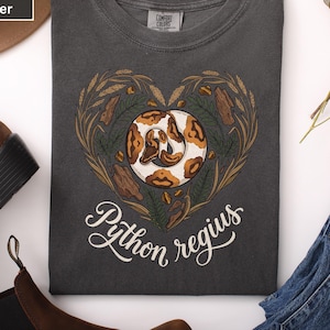 Op de afbeelding: Donkergrijs t-shirt met een hartvormig ontwerp met een slangenillustratie. De slang is wit en bruin, omringd door tarwe- en houtachtige elementen. De tekst "Python regius" is in cursieve letters geschreven.