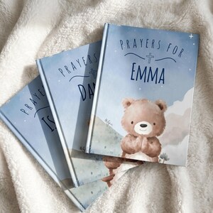 May include: Three personalized prayer books with blue covers. The books are titled "Prayers for Emma", "Prayers for Day", and "Prayers for Is". Each book features a teddy bear illustration and a cross symbol.