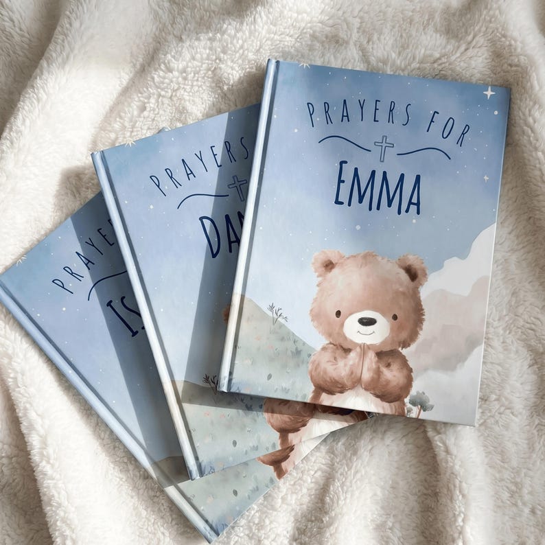 May include: Three personalized prayer books with blue covers. The books are titled "Prayers for Emma", "Prayers for Day", and "Prayers for Is". Each book features a teddy bear illustration and a cross symbol.