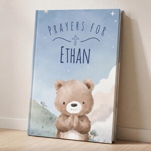 May include: A personalized children's book with a blue cover featuring the text "Prayers For Ethan" and a cute illustration of a teddy bear with its paws together. The background has a watercolor design with stars and mountains.