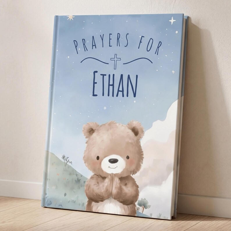 May include: A personalized children's book with a blue cover featuring the text "Prayers For Ethan" and a cute illustration of a teddy bear with its paws together. The background has a watercolor design with stars and mountains.