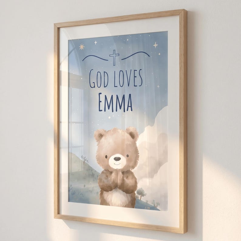 May include: Framed art print with a light wood frame. The artwork features a watercolor illustration of a teddy bear with its paws together, beneath the text "GOD LOVES EMMA" and a cross symbol, set against a blue sky background.