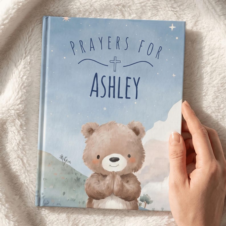 May include: A children's book with a blue cover featuring the text "Prayers for Ashley" and a cross. The cover also has an illustration of a teddy bear with its paws together in prayer. The background has a watercolor design with stars and mountains.