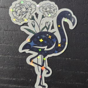 May include: A sticker featuring a stylized flamingo with a dark blue body and a constellation design. The flamingo holds two white flowers. The sticker has a holographic, iridescent effect.