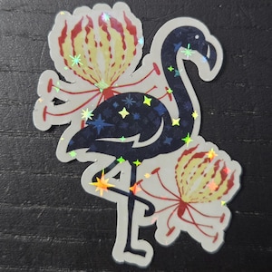 May include: A sticker featuring a stylized flamingo with a dark blue body and iridescent star accents. The flamingo is surrounded by two flowers with yellow and red petals. The sticker has a holographic finish.