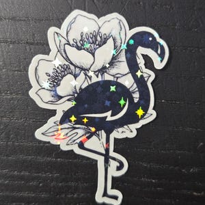 May include: A sticker featuring a dark blue flamingo with holographic star accents, standing on one leg. The flamingo is set against a backdrop of a detailed, black and white floral design.