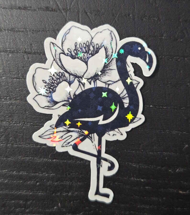 May include: A sticker featuring a dark blue flamingo with holographic star accents, standing on one leg. The flamingo is set against a backdrop of a detailed, black and white floral design.
