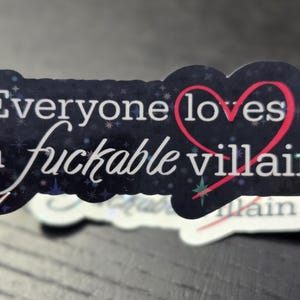 May include: A sticker with the text "Everyone loves a fuckable villain" in white script and a red heart. The sticker has a dark blue background with a starry pattern and a glittery finish.