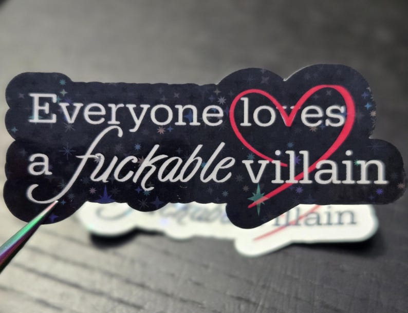 May include: A sticker with the text "Everyone loves a fuckable villain" in white script and a red heart. The sticker has a dark blue background with a starry pattern and a glittery finish.