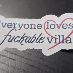 May include: A white sticker with the text "Everyone loves a fuckable villain" in black script and a red heart. The sticker has a holographic, iridescent sheen.