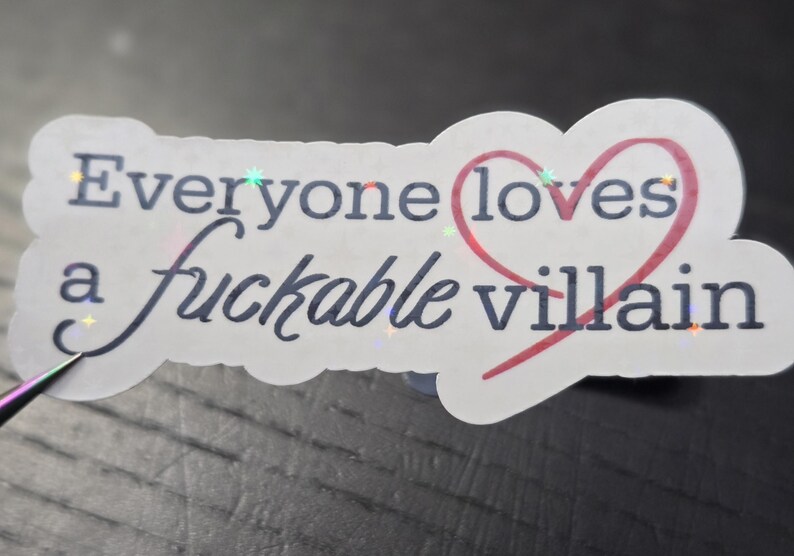 May include: A white sticker with the text "Everyone loves a fuckable villain" in black script and a red heart. The sticker has a holographic, iridescent sheen.