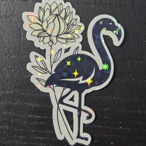 May include: A sticker featuring a stylized flamingo with a flower. The flamingo is dark blue with a starry pattern, and the flower is light green. The sticker has a holographic effect, with iridescent accents.