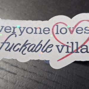 May include: A white sticker with the text "Everyone loves a fuckable villain" in dark blue, with a red heart around the word "loves". The sticker has a holographic effect.