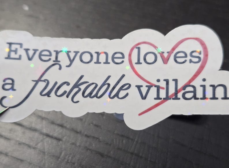 May include: A white sticker with the text "Everyone loves a fuckable villain" in dark blue, with a red heart around the word "loves". The sticker has a holographic effect.