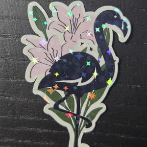 May include: A sticker featuring a stylized flamingo with a dark blue body and iridescent star-shaped accents. The flamingo is set against a backdrop of light pink flowers and green leaves, with a white border.