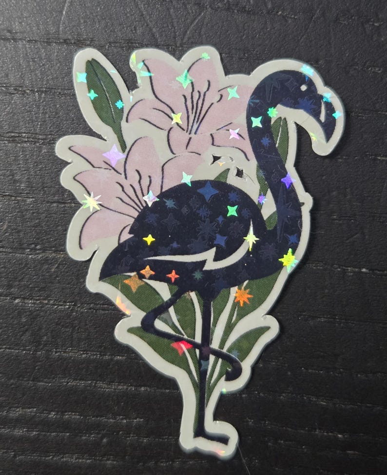 May include: A sticker featuring a stylized flamingo with a dark blue body and iridescent star-shaped accents. The flamingo is set against a backdrop of light pink flowers and green leaves, with a white border.