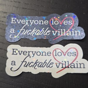May include: Two stickers with the phrase "Everyone loves a fuckable villain." One sticker is on a dark blue background with a star pattern, and the other is on a white background. Both have a pink heart.