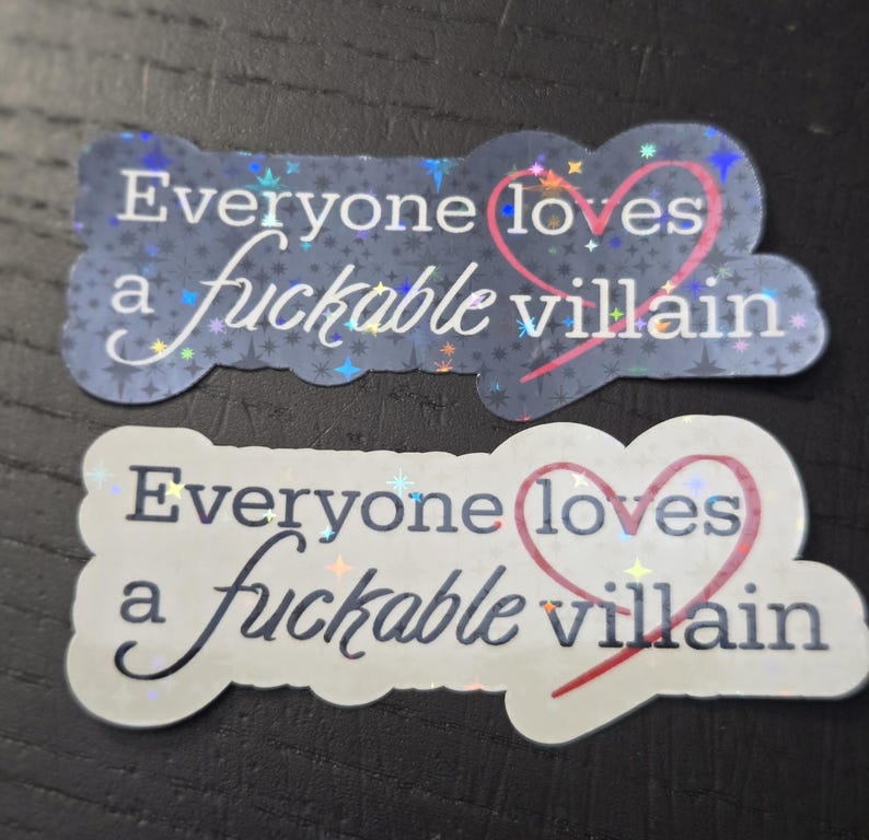 May include: Two stickers with the phrase "Everyone loves a fuckable villain." One sticker is on a dark blue background with a star pattern, and the other is on a white background. Both have a pink heart.