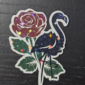 May include: A holographic sticker featuring a flamingo and a rose. The flamingo is dark blue with a starry pattern, and the rose is burgundy with holographic accents. Green leaves surround the flowers and bird.