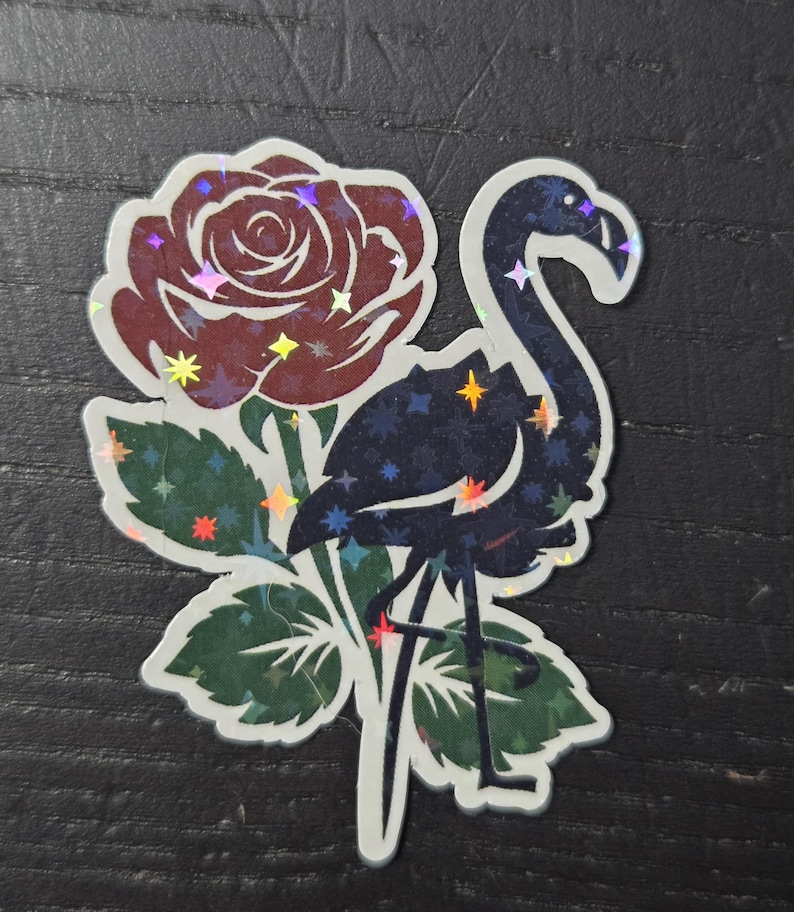 May include: A holographic sticker featuring a flamingo and a rose. The flamingo is dark blue with a starry pattern, and the rose is burgundy with holographic accents. Green leaves surround the flowers and bird.
