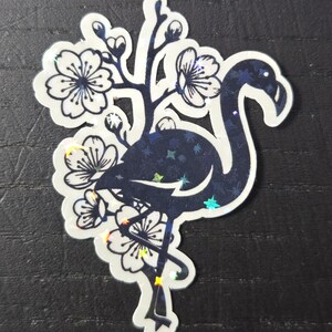 May include: A die-cut sticker featuring a stylized flamingo silhouette in dark blue with a starry pattern. The flamingo is surrounded by white cherry blossoms with dark blue outlines. The sticker has a holographic, iridescent finish.