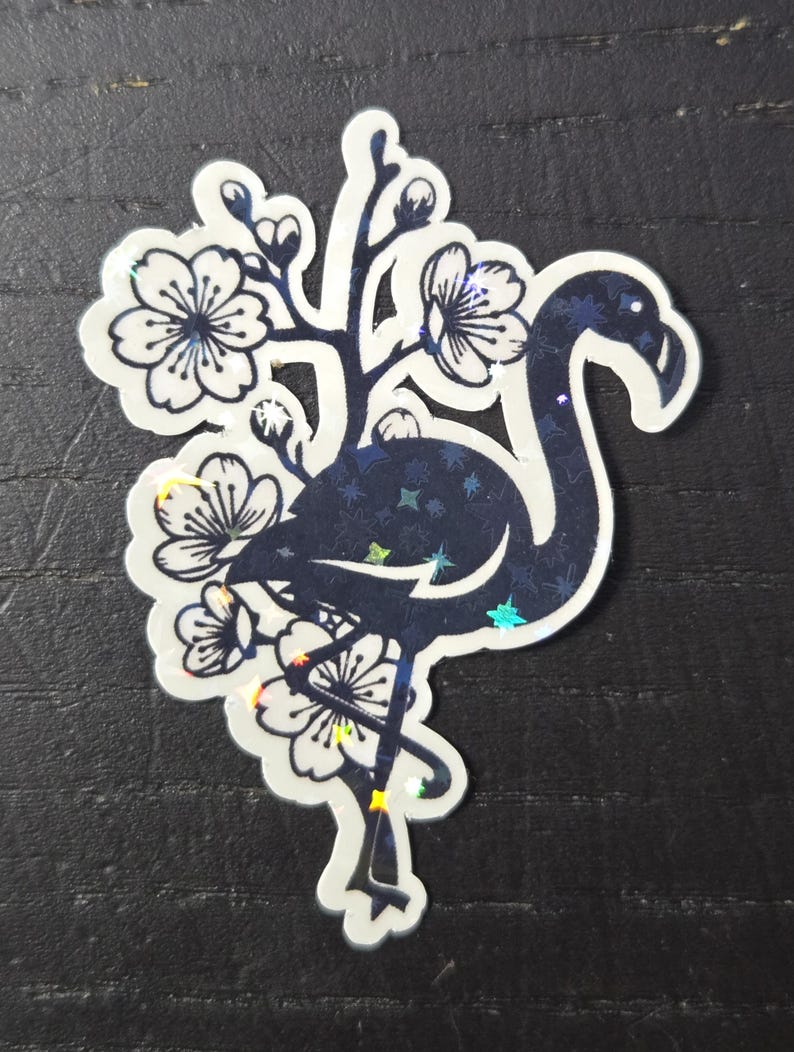 May include: A die-cut sticker featuring a stylized flamingo silhouette in dark blue with a starry pattern. The flamingo is surrounded by white cherry blossoms with dark blue outlines. The sticker has a holographic, iridescent finish.