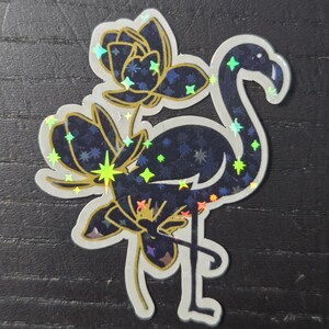 May include: A holographic sticker featuring a flamingo and floral design. The flamingo is dark blue with a starry pattern, and the flowers are gold and dark blue. The sticker has a white border and is on a dark background.