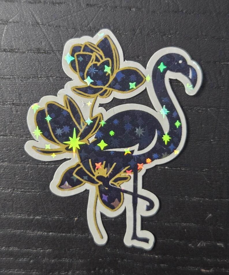 May include: A holographic sticker featuring a flamingo and floral design. The flamingo is dark blue with a starry pattern, and the flowers are gold and dark blue. The sticker has a white border and is on a dark background.