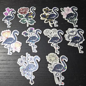 May include: A collection of flamingo stickers with floral accents. Each sticker features a dark blue flamingo with various flower designs, including roses, chrysanthemums, and other blooms. The stickers have a holographic effect and are arranged on a dark surface.