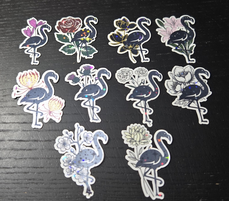 May include: A collection of flamingo stickers with floral accents. Each sticker features a dark blue flamingo with various flower designs, including roses, chrysanthemums, and other blooms. The stickers have a holographic effect and are arranged on a dark surface.