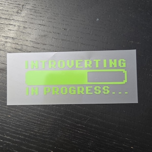 May include: A rectangular sticker with a translucent backing featuring neon green pixelated text that reads "INTROVERTING IN PROGRESS..." above a partially filled neon green loading bar graphic, designed with a retro computer interface aesthetic.
