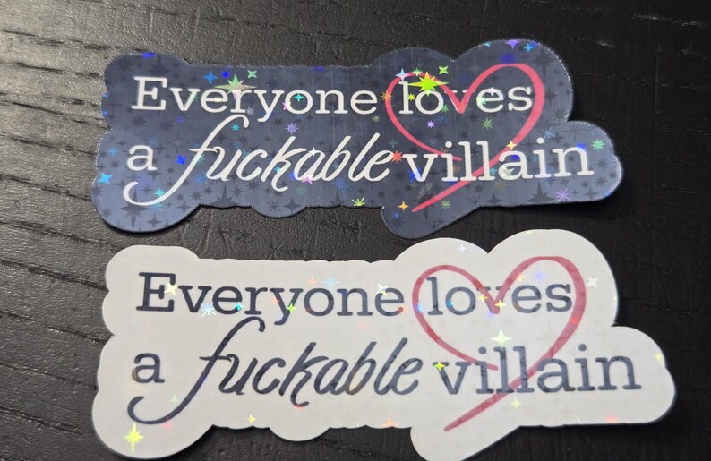 May include: Two stickers with the phrase "Everyone loves a fuckable villain." One sticker is dark blue with a starry background, the other is white. Both have a pink heart.