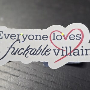 May include: A white sticker with the text "Everyone loves a fuckable villain" in black script and a red heart. The sticker has a scalloped edge and is on a dark surface.