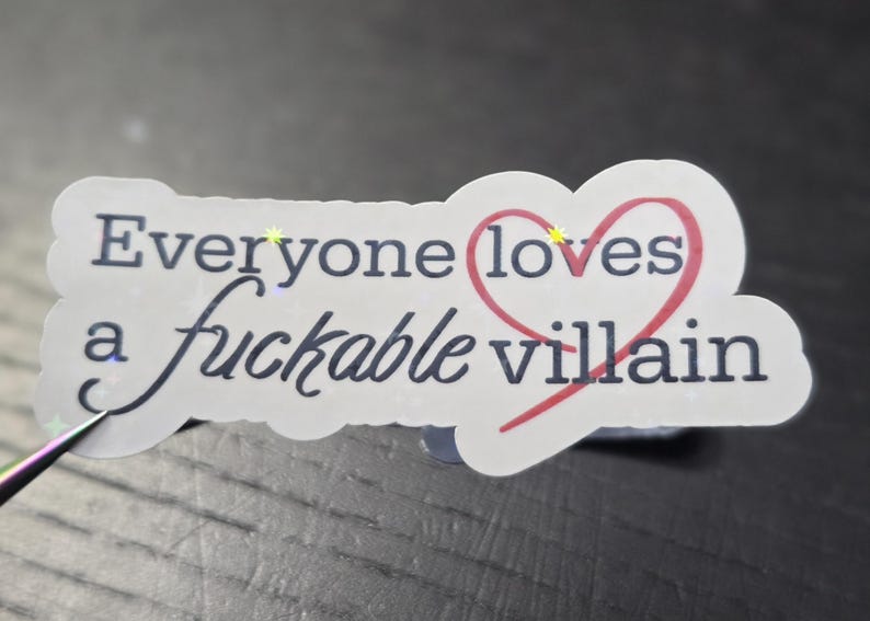 May include: A white sticker with the text "Everyone loves a fuckable villain" in black script and a red heart. The sticker has a scalloped edge and is on a dark surface.