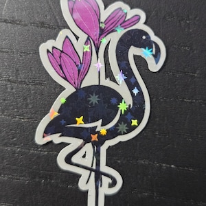 May include: A sticker featuring a stylized flamingo with a dark blue body and iridescent accents. The flamingo is adorned with purple flower blossoms and colorful star-shaped details. The sticker has a white border.
