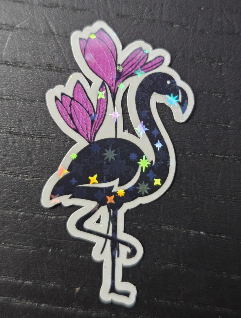May include: A sticker featuring a stylized flamingo with a dark blue body and iridescent accents. The flamingo is adorned with purple flower blossoms and colorful star-shaped details. The sticker has a white border.