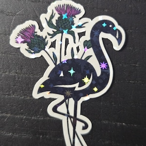 May include: A holographic sticker featuring a flamingo with a dark blue body and a white outline. The flamingo is adorned with star-shaped accents and is paired with purple thistle flowers.