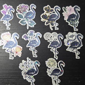 May include: A collection of ten flamingo stickers with floral accents. Each sticker features a dark blue flamingo with a shimmering, iridescent finish, paired with various flower designs in shades of pink, purple, and gold. The stickers are on a dark background.