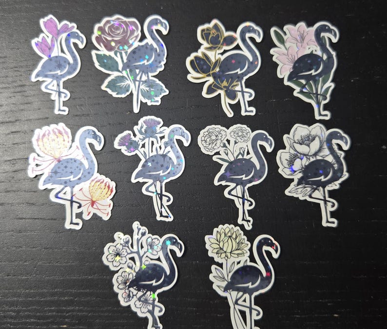 May include: A collection of ten flamingo stickers with floral accents. Each sticker features a dark blue flamingo with a shimmering, iridescent finish, paired with various flower designs in shades of pink, purple, and gold. The stickers are on a dark background.