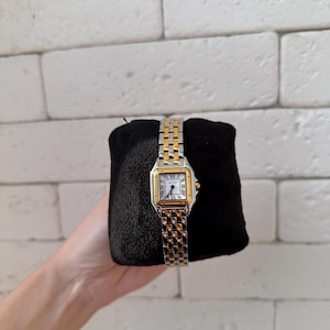 May include: A gold and silver wristwatch with a square face and Roman numeral hour markers. The watch has a two-tone metal band and is displayed on a black surface. The watch face is white.