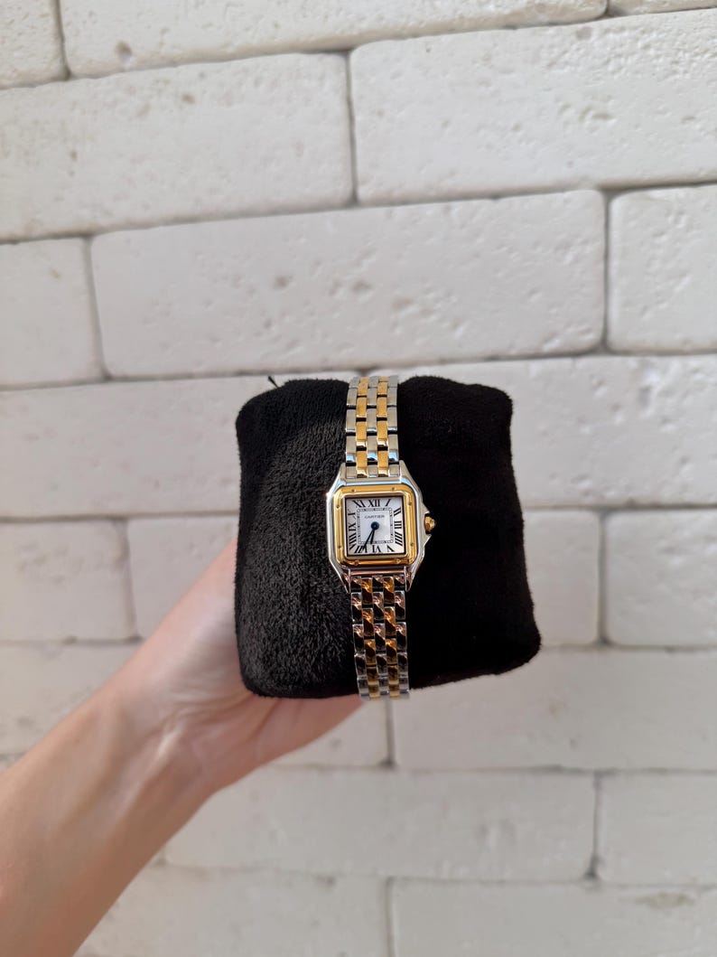 May include: A gold and silver wristwatch with a square face and Roman numeral hour markers. The watch has a two-tone metal band and is displayed on a black surface. The watch face is white.